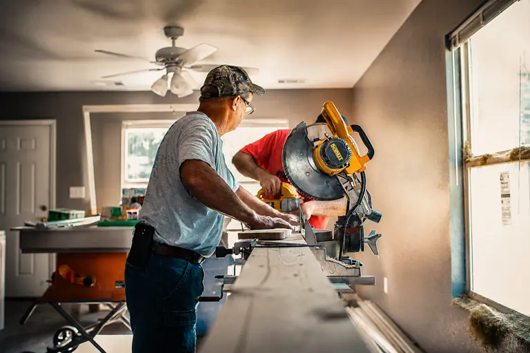 Two contractors using a miter saw to cut trim inside a home under renovation